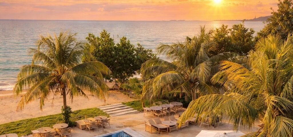 Sunset over the ocean seen from a tropical resort, with palm trees, a blue rectangular pool, and lounge seating nearby.