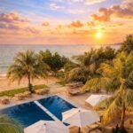 Sunset over the ocean seen from a tropical resort, with palm trees, a blue rectangular pool, and lounge seating nearby.