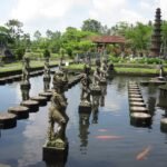 Stone statues rising from a reflective pond, lined by square stepping stones in a lush temple garden.