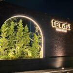 Night scene of an illuminated restaurant sign reading 'terata' with 'SOCIAL DINING' on a brick wall beside a lit circular plant display.