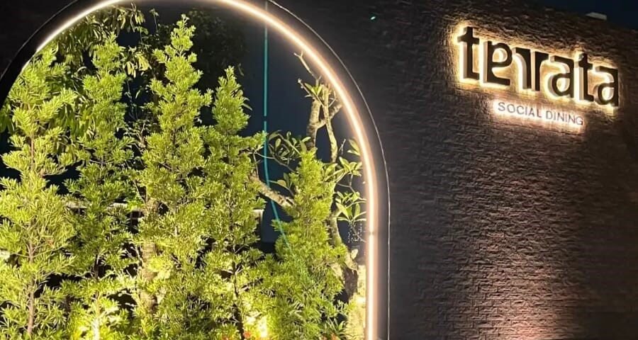 Night scene of an illuminated restaurant sign reading 'terata' with 'SOCIAL DINING' on a brick wall beside a lit circular plant display.