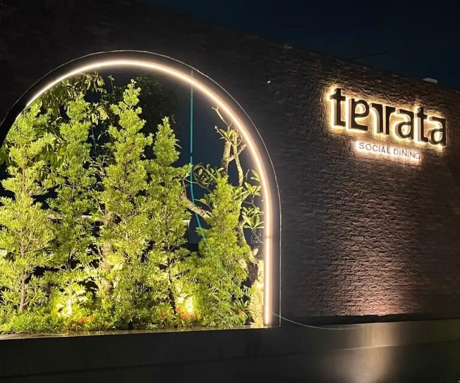 Night scene of an illuminated restaurant sign reading 'terata' with 'SOCIAL DINING' on a brick wall beside a lit circular plant display.