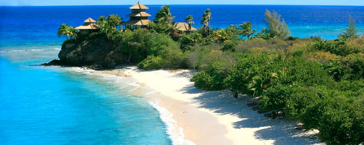 Tropical beach with white sand, turquoise water, and palm trees on a rocky headland with thatched huts overlooking the ocean.