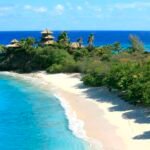 Tropical beach with white sand, turquoise water, and palm trees on a rocky headland with thatched huts overlooking the ocean.