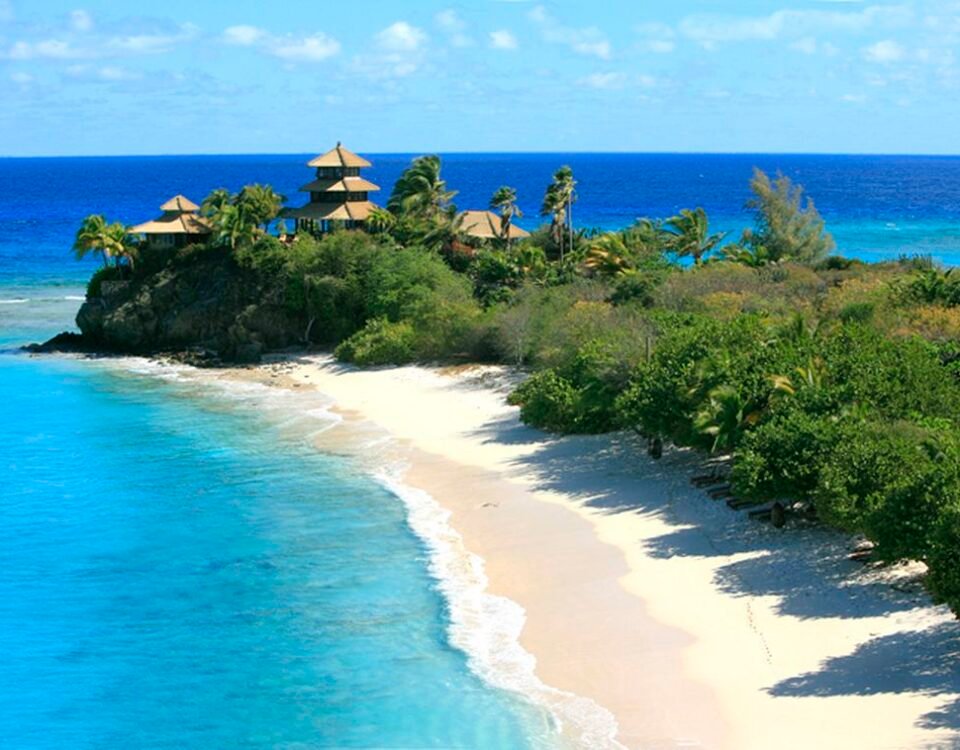 Tropical beach with white sand, turquoise water, and palm trees on a rocky headland with thatched huts overlooking the ocean.