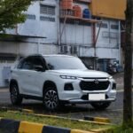 White SUV parked on a city street with a building and orange water tanks on a raised platform in the background.