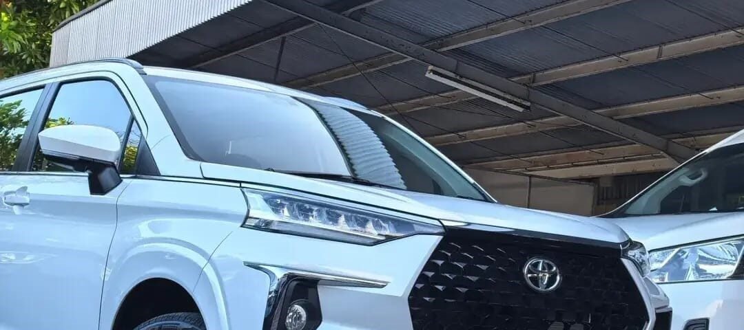 White Toyota Veloz Hybrid EV parked under a metal carport, front grille and emblem visible, glossy alloy wheels.