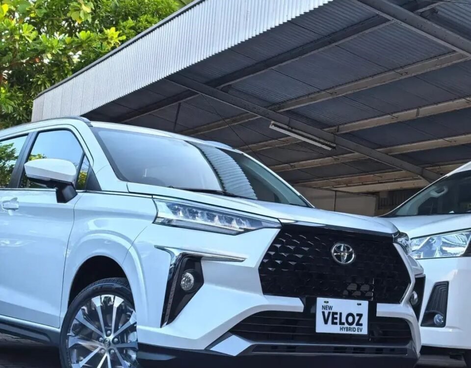 White Toyota Veloz Hybrid EV parked under a metal carport, front grille and emblem visible, glossy alloy wheels.