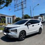 White Mitsubishi SUV parked on a sunny urban street with buildings, a flagpole, and streetlights in the background.
