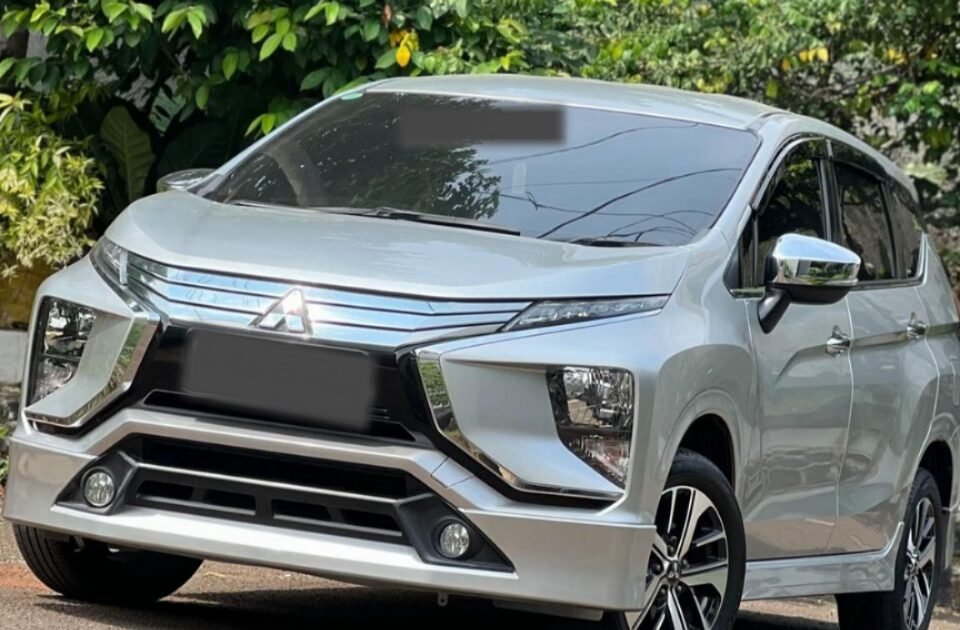 Silver Mitsubishi SUV parked on a street beside lush green tropical plants and a concrete wall.
