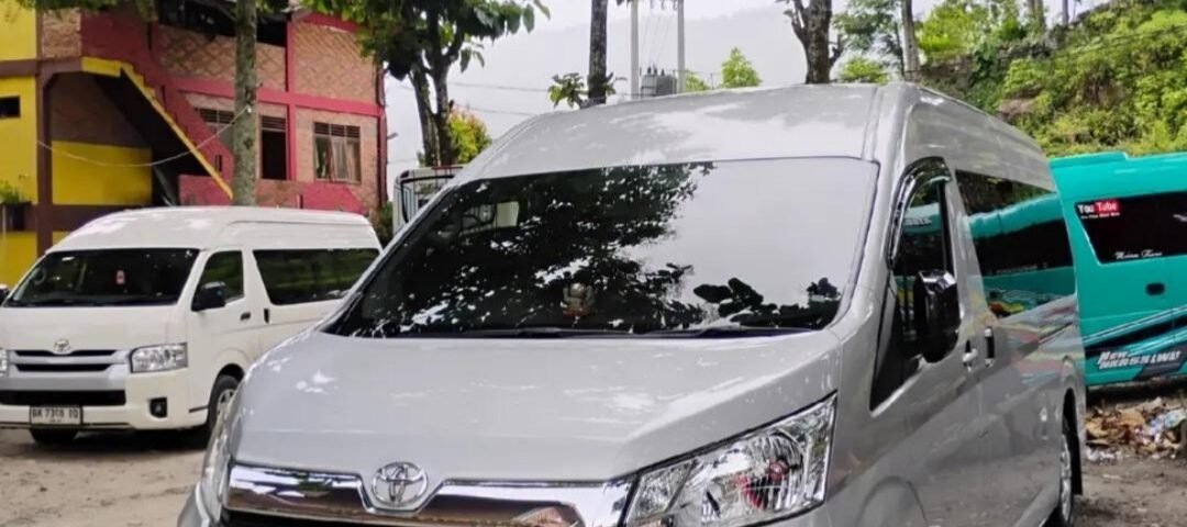 Front three-quarter view of a white Toyota van parked in a lot, with trees overhead and other vans in the background.