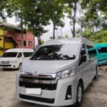 Front three-quarter view of a white Toyota van parked in a lot, with trees overhead and other vans in the background.