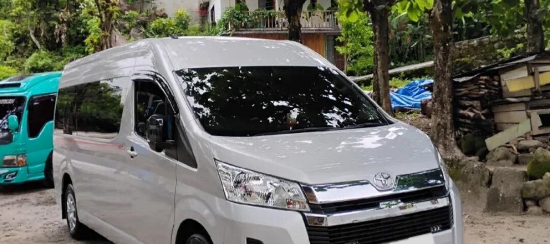 White Toyota van parked on a dirt lot under leafy trees, with a turquoise van and houses in the background.