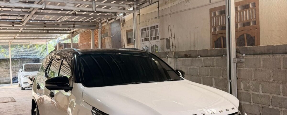 White Toyota Fortuner SUV parked in a metal-framed carport under a corrugated metal roof in a driveway.