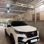 White Toyota Fortuner SUV parked in a metal-framed carport under a corrugated metal roof in a driveway.