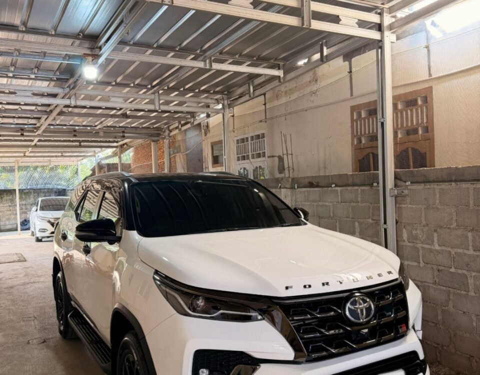 White Toyota Fortuner SUV parked in a metal-framed carport under a corrugated metal roof in a driveway.