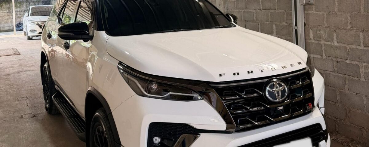 White Toyota Fortuner SUV parked in a metal-framed carport under a corrugated metal roof in a driveway.