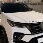 White Toyota Fortuner SUV parked in a metal-framed carport under a corrugated metal roof in a driveway.