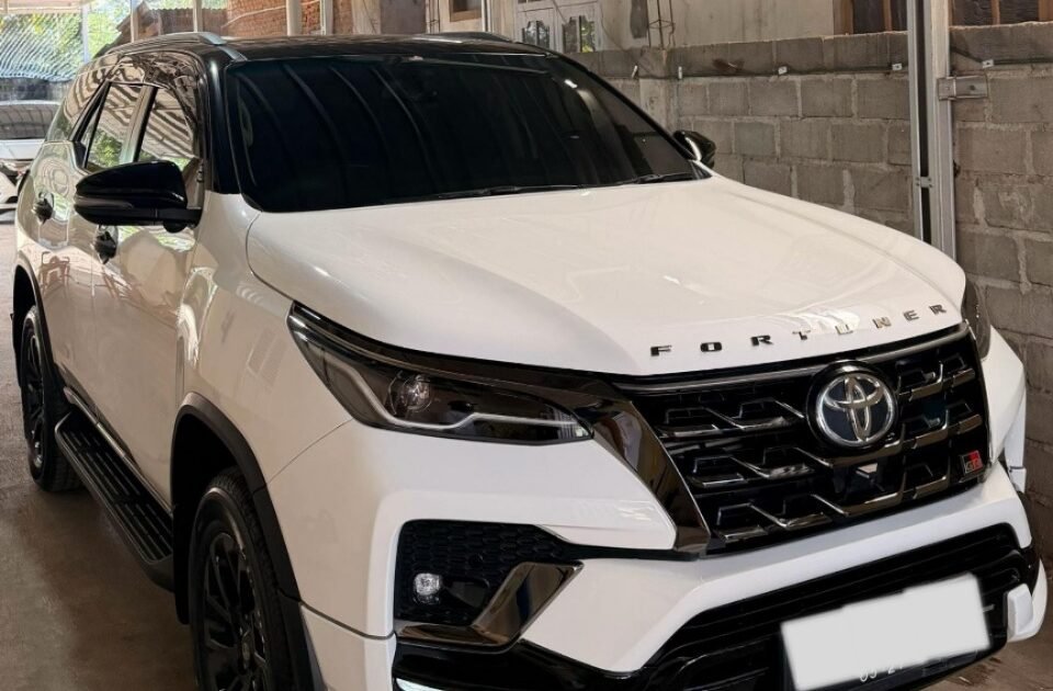 White Toyota Fortuner SUV parked in a metal-framed carport under a corrugated metal roof in a driveway.