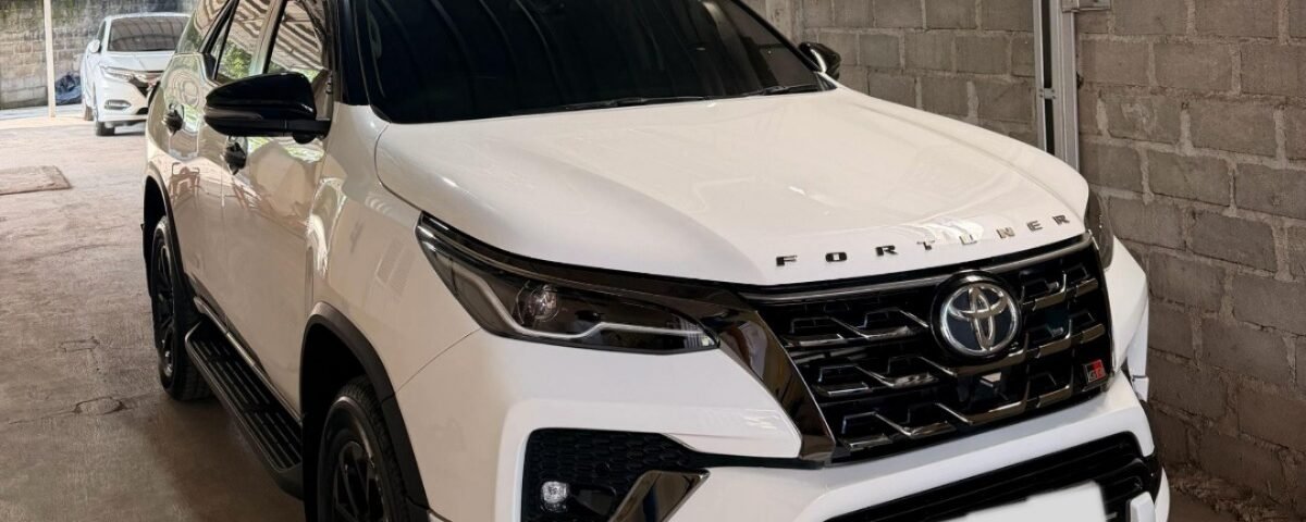 White Toyota Fortuner SUV parked in a covered garage beside a brick wall, front three-quarter view with tinted windows and black trim.