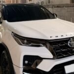 White Toyota Fortuner SUV parked in a covered garage beside a brick wall, front three-quarter view with tinted windows and black trim.