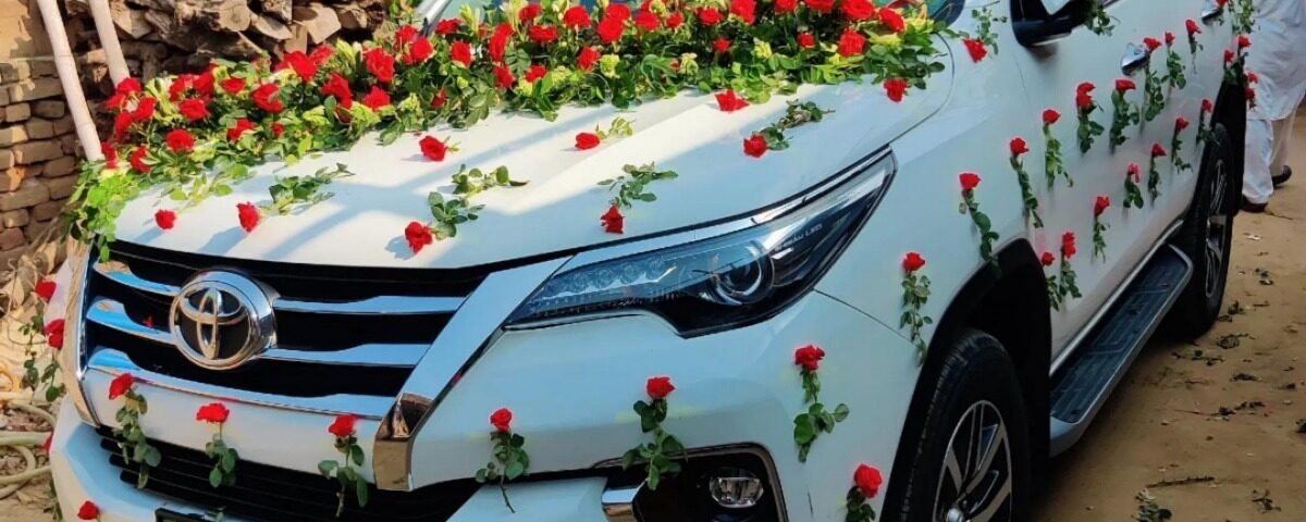 White Toyota SUV decorated with red roses and green garlands for a wedding, parked in a dusty yard with wood stacks behind it.