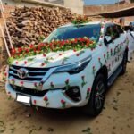 White Toyota SUV decorated with red roses and green garlands for a wedding, parked in a dusty yard with wood stacks behind it.