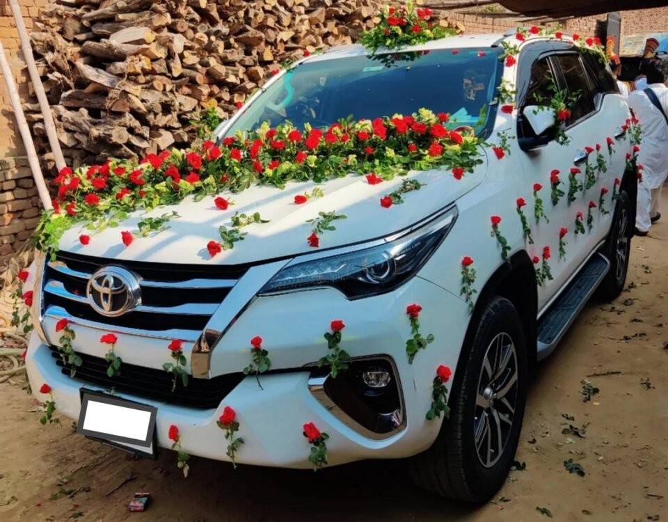 White Toyota SUV decorated with red roses and green garlands for a wedding, parked in a dusty yard with wood stacks behind it.