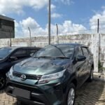Dark gray SUV parked in a lot beside a weathered concrete wall with barbed wire, under a blue sky with fluffy clouds.