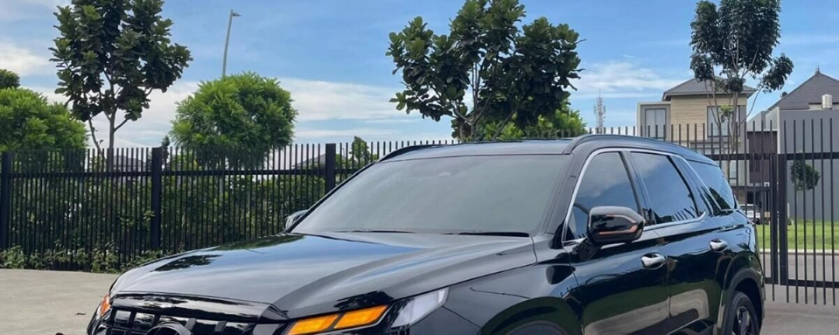 Black SUV parked on a paved lot with a metal fence, trees, and a blue sky behind it.