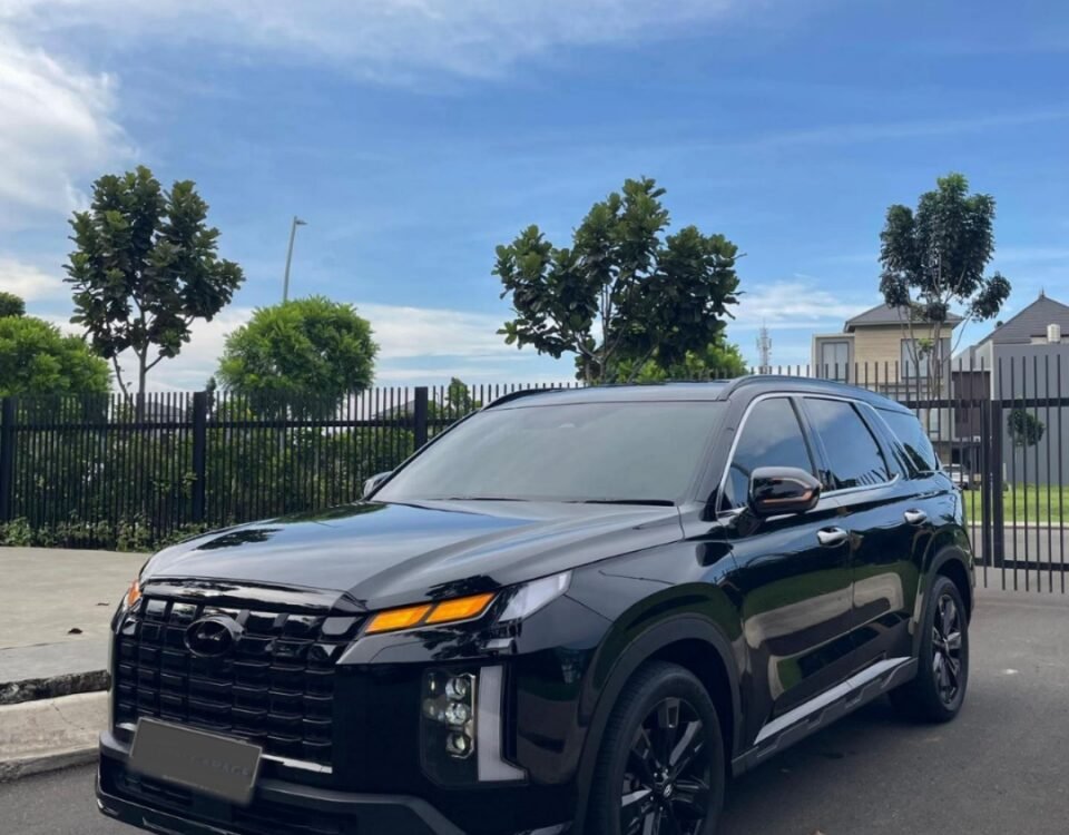 Black SUV parked on a paved lot with a metal fence, trees, and a blue sky behind it.
