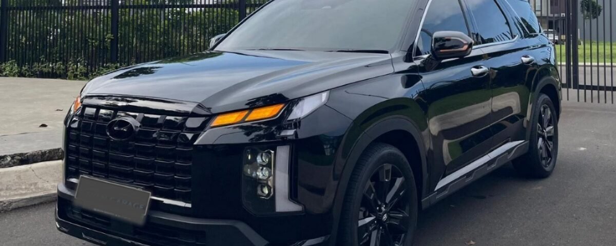 Black SUV parked on a paved lot with a metal fence, trees, and a blue sky behind it.