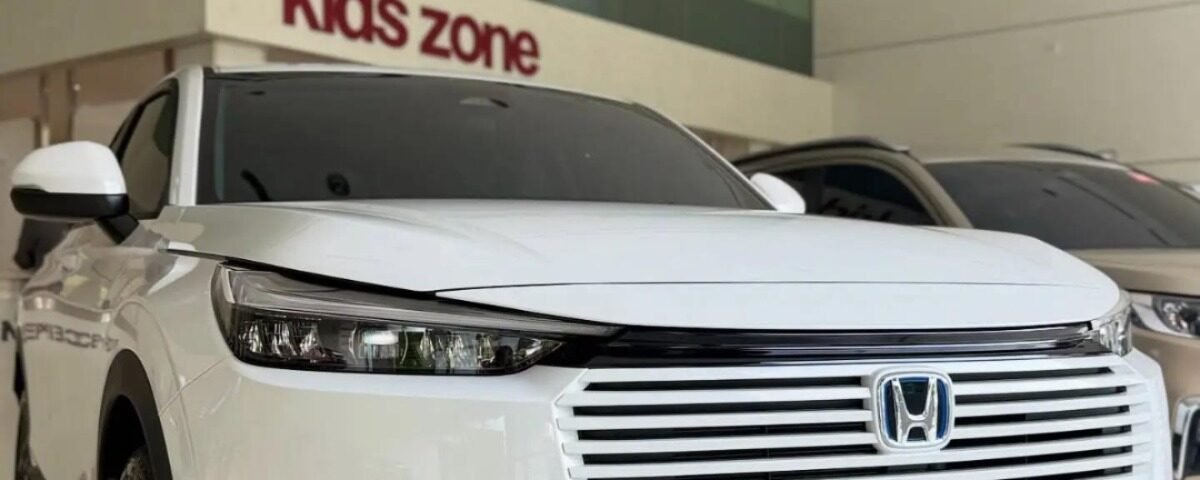 White Honda SUV on a showroom floor, front view with grille and logo visible, glass wall with 'kids zone' sign behind.