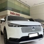 White Honda SUV on a showroom floor, front view with grille and logo visible, glass wall with 'kids zone' sign behind.