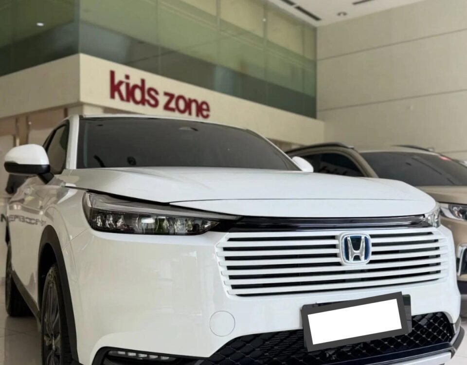 White Honda SUV on a showroom floor, front view with grille and logo visible, glass wall with 'kids zone' sign behind.