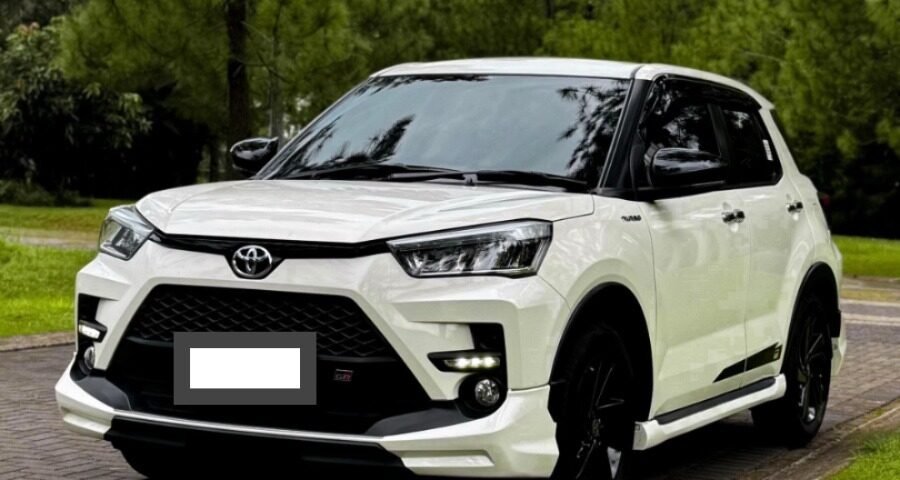 White Toyota SUV parked on a brick driveway beside a green lawn with tall pine trees in the background.