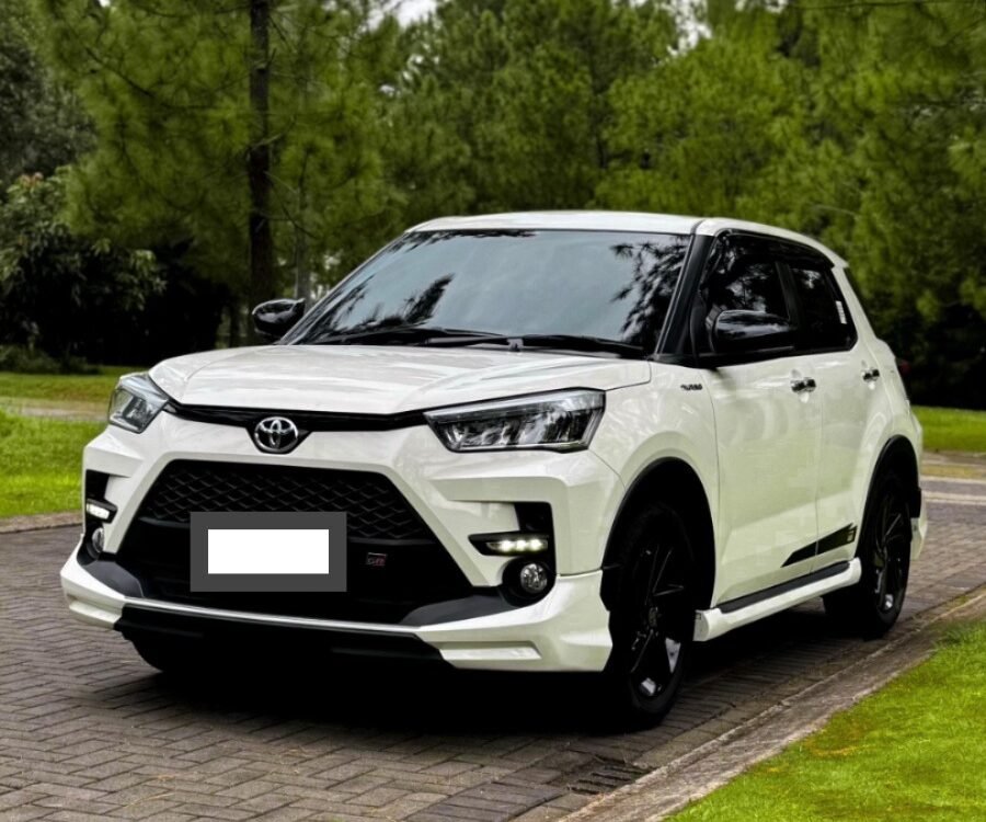 White Toyota SUV parked on a brick driveway beside a green lawn with tall pine trees in the background.