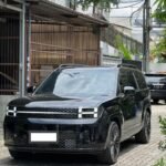 Front three-quarter view of a black SUV parked on a brick driveway with a gated building in the background and overhead power lines.