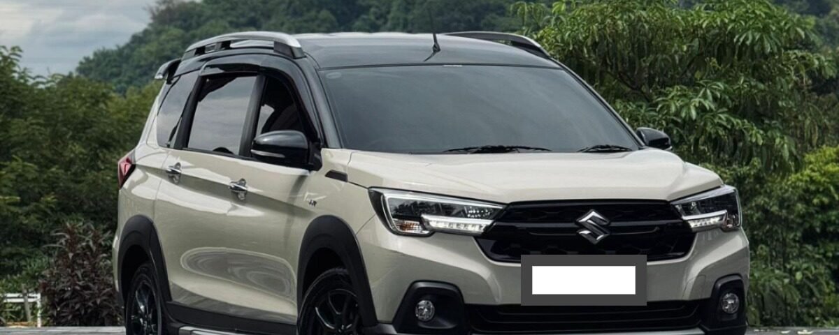 Beige Suzuki SUV parked on a checkered driveway with green hills in the background, front three-quarter view with roof rails and black wheels.