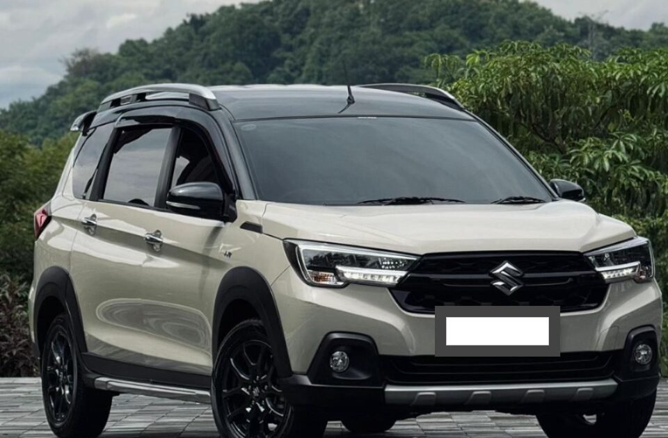 Beige Suzuki SUV parked on a checkered driveway with green hills in the background, front three-quarter view with roof rails and black wheels.