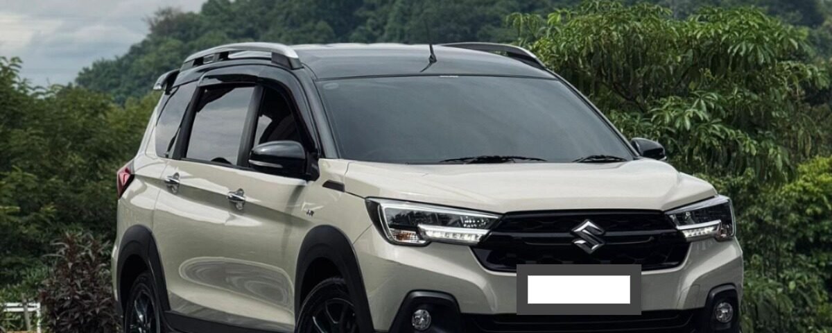 Beige Suzuki SUV in a three-quarter front view parked on a checkered pavement with green hills and cloudy sky behind.