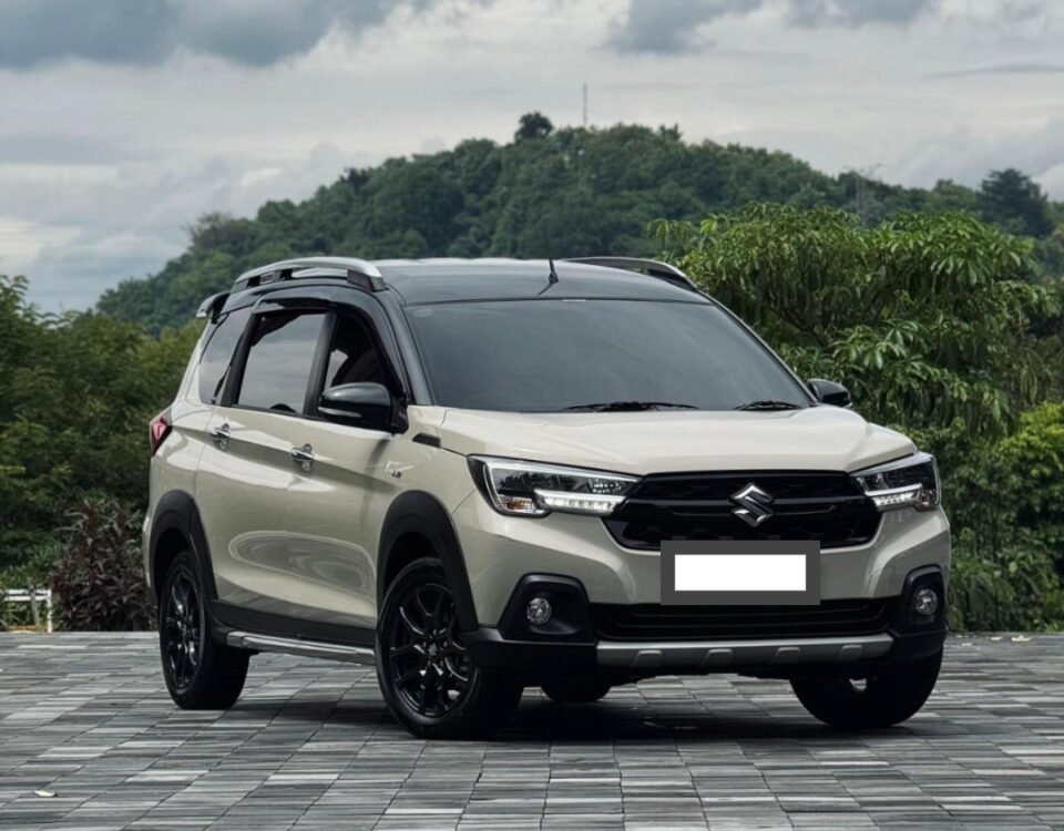 Beige Suzuki SUV in a three-quarter front view parked on a checkered pavement with green hills and cloudy sky behind.