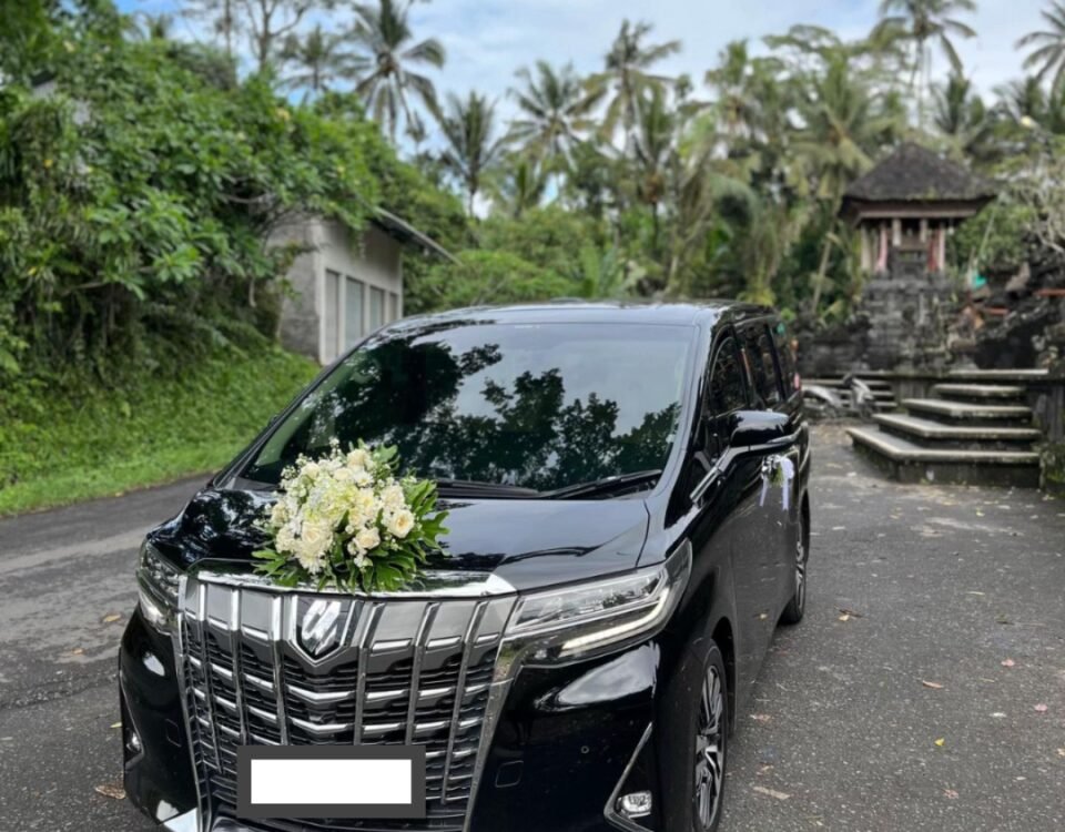 Black wedding car with a white flower bouquet on its hood, parked on a tropical road with palm trees nearby.