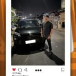 Man in a black shirt posing beside a black SUV on a nighttime city street and storefronts in the background, group photo-style post context included.