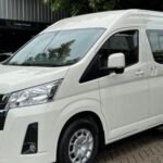 White Toyota high-roof passenger van parked in a brick-paved lot with green trees behind it.