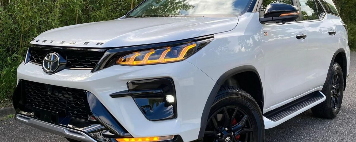 White Toyota SUV parked on a paved surface with dense green bushes and trees in the background.