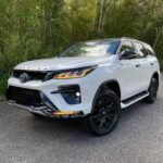 White Toyota SUV parked on a paved surface with dense green bushes and trees in the background.
