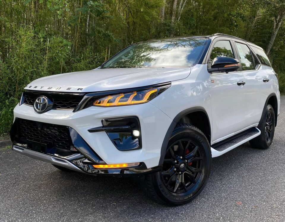 White Toyota SUV parked on a paved surface with dense green bushes and trees in the background.