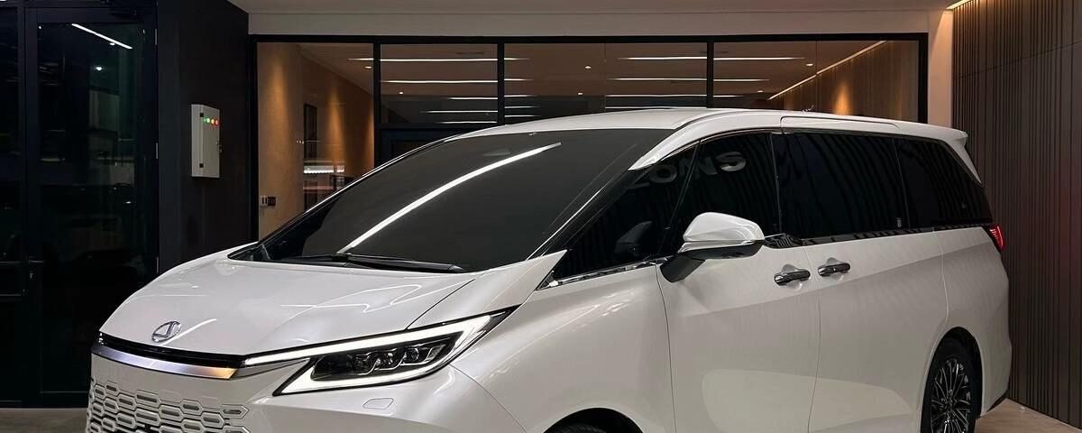 White luxury minivan parked in a modern showroom, featuring a bold front grille and dark-tinted windows under bright overhead lighting.