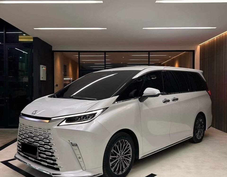 White luxury minivan parked in a modern showroom, featuring a bold front grille and dark-tinted windows under bright overhead lighting.